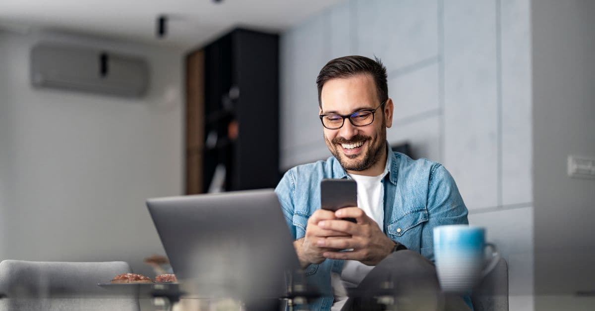 Homem sorridente usando o celular diante do notebook