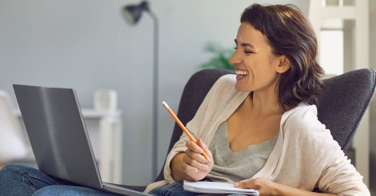 Mulher sorridente fazendo anotações enquanto assiste a uma aula no notebook