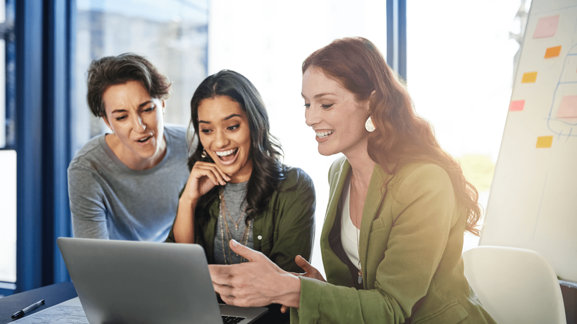 Três colegas sorridentes reunidas em torno de um notebook no escritório