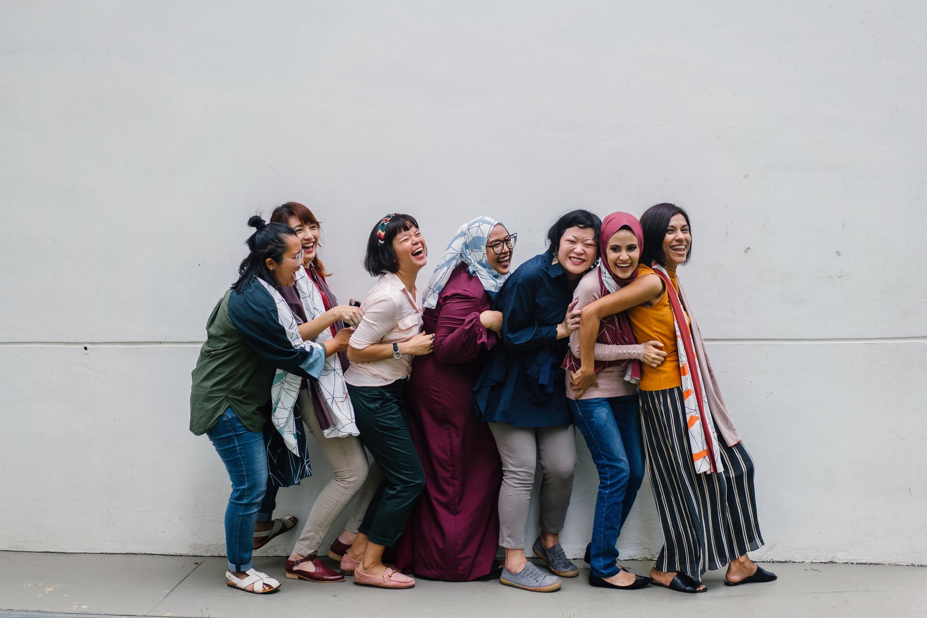 Grupo de mulheres de diferentes origens rindo e abraçadas em frente a uma parede branca